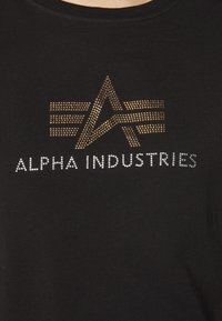 T-shirt en coton noir avec un logo en strass dorés et argentés portant la mention "ALPHA INDUSTRIES" accompagné d'un design d'étoile stylisé.