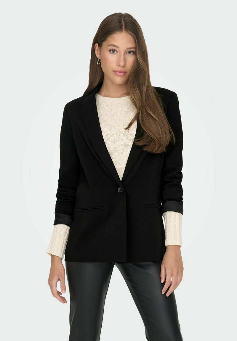 Chaqueta negra ajustada con solapas de muesca y un solo botón. Lleva puesta una chaqueta de punto grueso color crema, combinada con pantalones de cuero sintético negros.