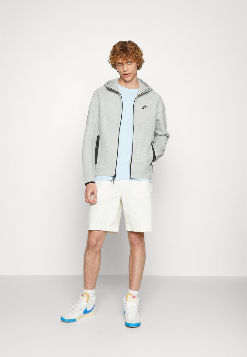 Sudadera gris con cremallera y acentos negros, combinada con una camiseta de color azul claro y pantalones cortos blancos. El modelo lleva zapatillas blancas con detalles azules.