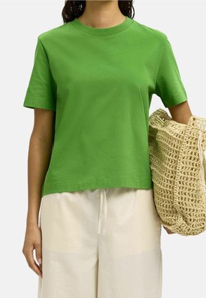 Personne portant un t-shirt vert uni à manches courtes et un pantalon beige clair, tenant un grand sac tressé beige à leur côté.