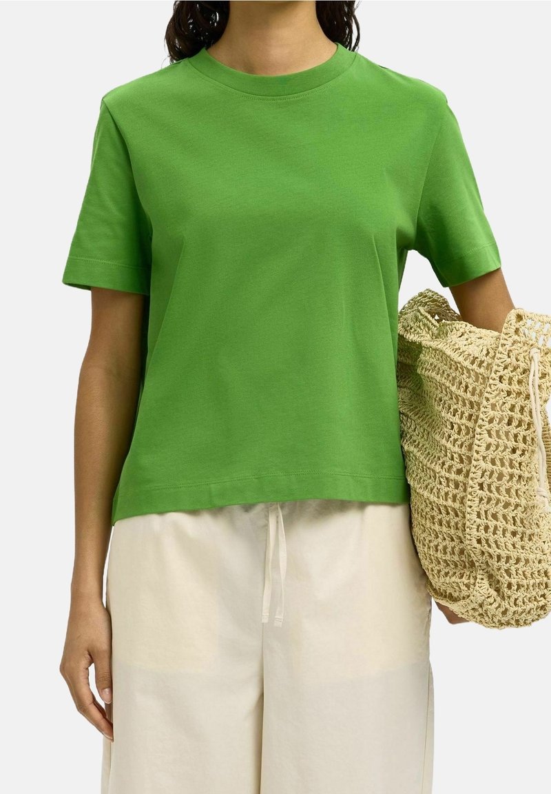 Personne portant un t-shirt vert uni à manches courtes et un pantalon beige clair, tenant un grand sac tressé beige à leur côté.
