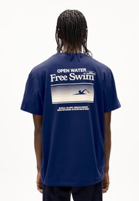 T-shirt en coton bleu marine avec un graphisme blanc au dos, portant le texte "NAGE LIBRE ENEAU OUVERTE" et une illustration d'un nageur.