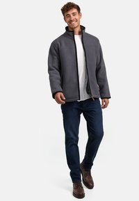 Chaqueta de forro polar gris con un cuello alto, cierre de cremallera y ribete negro, combinada con jeans oscuros y botas de cordones marrones.