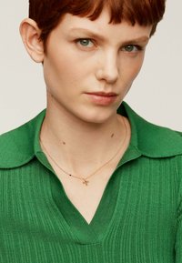Jersey de punto acanalado verde con cuello en V, acompañado de un delicado collar de oro con pequeños acentos decorativos.