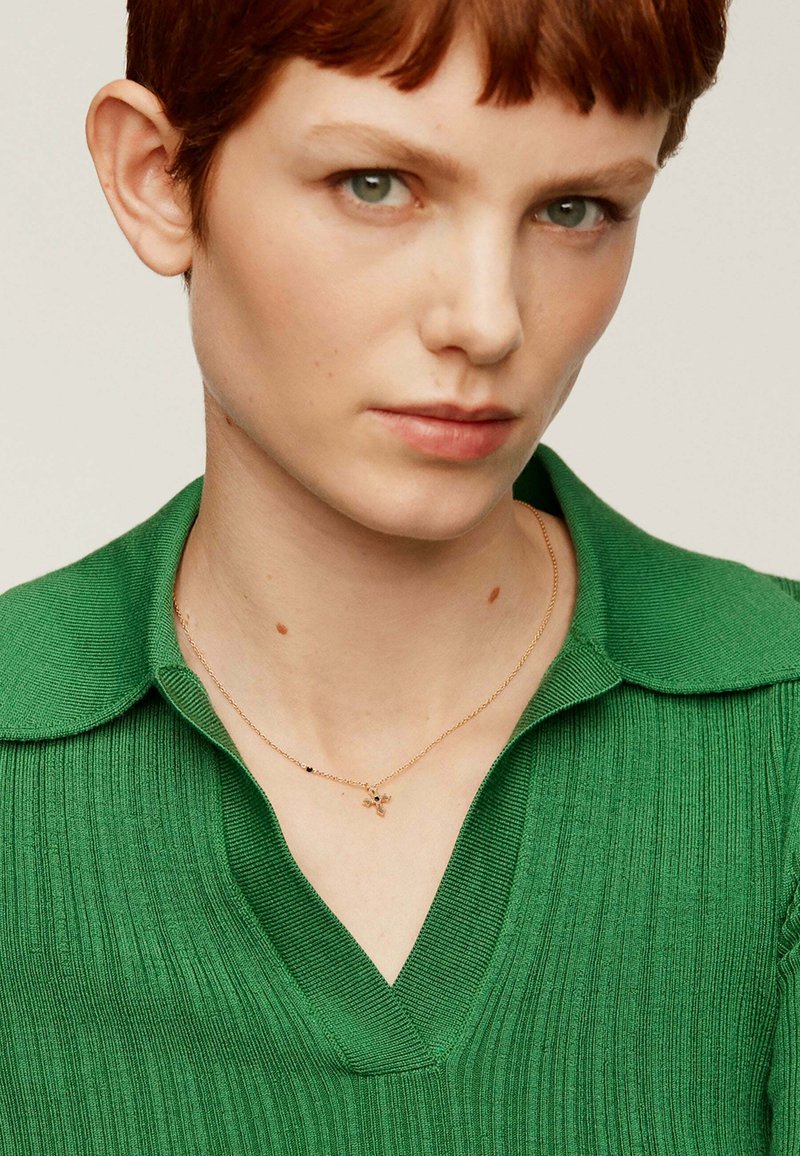 Jersey de punto acanalado verde con cuello en V, acompañado de un delicado collar de oro con pequeños acentos decorativos.