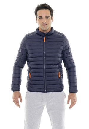 Un homme se tient devant un fond blanc, vêtu d’une veste matelassée bleu marine avec des tirettes de fermeture éclair orange et d’un pantalon gris clair.