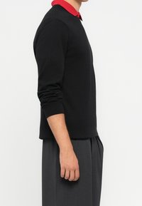 Homme portant un pull noir à manches longues par-dessus une chemise rouge à col, avec un pantalon gris plissé, debout de profil sur un fond uni.