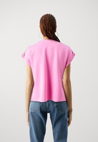 Persona con trenzas largas que lleva pendientes de aro, una camiseta rosa de manga corta con puños enrollados y jeans azules, de pie frente a un fondo liso.