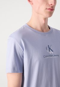 Camiseta de algodón de color púrpura claro con mangas cortas, con un logotipo de la marca impreso en colores contrastantes en la parte frontal. Textura suave y corte clásico.