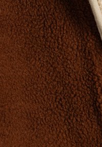 Tela marrón y esponjosa con una superficie suave y texturizada. El borde presenta una tela lisa de color crema. En general, tiene una apariencia cálida y acogedora.