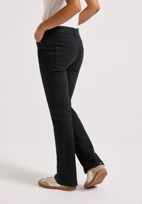 Jean flare noir en tissu mélangé de coton, avec poches arrière et taille haute. Associés à des baskets de couleur claire.