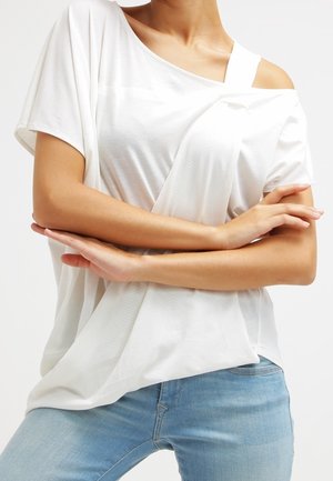 Osoba ubrana w luźną białą bluzkę z odkrytymi ramionami i jasnoniebieskie dżinsy, z rękami skrzyżowanymi na wysokości talii, na jednolitym tle.