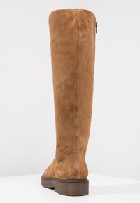 Höga bruna mockaboots för kvinnor med sidodragkedja och låg staplad sula visade bakifrån på en vit yta.