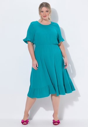 Frau in einem tealfarbenen knielangen Kleid mit Rüschen an Ärmeln und Saum, trägt leuchtend pinke offene Schuhe mit Schleifen und steht vor einem weißen Hintergrund.