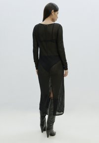 Schwarzes, langärmeliges, transparentes Strickkleid mit Seitenschlitzen und strukturiertem Stoff. Kombiniert mit kniehohen, spitz zulaufenden schwarzen Stiefeln.