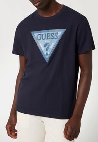 Marineblauwe katoenen t-shirt met een driehoekige denim patch met het "GUESS" logo en uitgerafelde randen. Ronde halslijn en korte mouwen.
