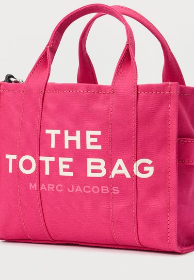 Ροζ καμβαδένια τσάντα ώμου με δύο κοντά χερούλια. Φέρει έντονη λευκή γραφή με τα λόγια "THE TOTE BAG" και "MARC JACOBS." Απλός, δομημένος σχεδιασμός.
