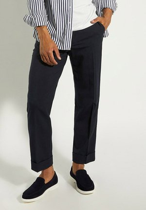 Pantalons bleu marine à coupe ajustée, avec des ourlets roulés. Associés à des chaussures en daim foncé à enfiler et une chemise rayée.