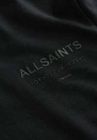 Musta kangas, jossa on kohokuvioitu teksti "ALLSAINTS" ja japaninkielisiä merkkejä, ja "LONDON" pienemmällä fontilla alla. Pehmeä, teksturoitu pinta.