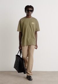 Oliven grøn oversized t-shirt med logo, beige lige bukser, sort taske, solbriller og sorte sko. Ensfarvet baggrund.