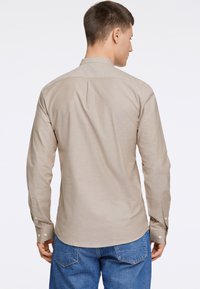 Camisa de manga larga color beige claro con cuello abotonado, con un pliegue en la espalda y puños de botón. Llevada con jeans de mezclilla azules.