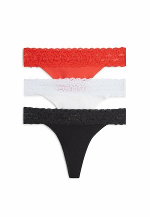 Giovane donna in piedi a piedi nudi che indossa un top bianco senza maniche e biancheria intima nera con bordi in pizzo su uno sfondo bianco semplice.