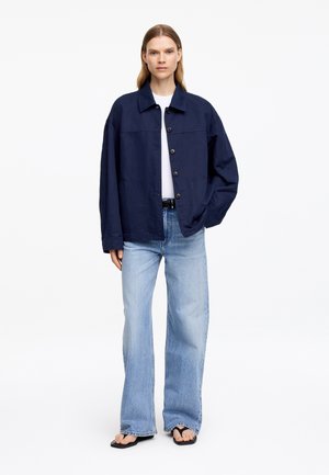 Femme portant une veste bleu marine boutonnée, un t-shirt blanc, un jean large bleu clair, une ceinture noire et des tongs noires, debout avec une main dans la poche.