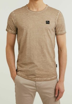 Mężczyzna w jasnobrązowej koszulce z krótkim rękawem z małym czarnym logo na piersi oraz beżowych spodniach, z rękami w kieszeniach.