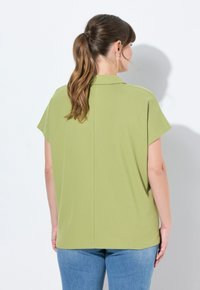 Top a maniche corte di colore verde chiaro, realizzato in tessuto strutturato, presenta una linea dritta e un dettaglio di cucitura posteriori, abbinato a jeans blu.
