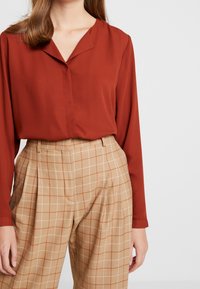 Rust long-sleeve blouse with a V-neckline paired with high-waisted, beige plaid trousers featuring wide pleats and a checked pattern.