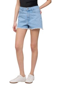 Shorts in denim blu chiaro con orlo sfrangiato, tasche frontali e accenti di borchie sottili. Abbinati a scarpe sportive bianche.