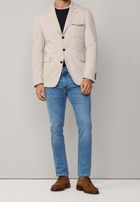 Beige Baumwollblazer mit einer Brusttasche, zwei Fronttaschen und schwarzen Knöpfen, kombiniert mit hellblauen Jeans und braunen Wildleder-Schuhen.