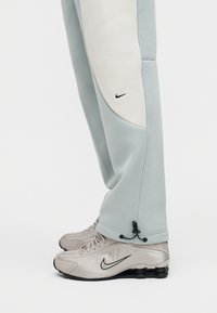 Πόδι σε ανοιχτό γκρι φόρμα με λευκή επιφάνεια και μαύρο λογότυπο Nike, φορώντας μπεζ αθλητικά παπούτσια Nike με μαύρη σόλα και λογότυπο swoosh.