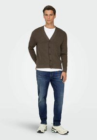 Brun cardigan med V-hals, knapper foran, kombinert med en hvit skjorte, blå jeans og hvite joggesko med grønne detaljer.