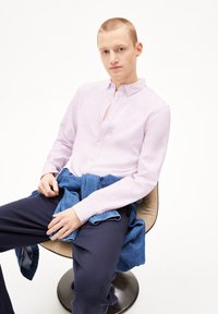 Lichtpaars overhemd met lange mouwen, gecombineerd met donkere broek. Denim trui over de knieën gedrapeerd, zittend op een gebogen stoel.