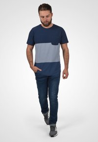 Tamno plavi T-shirt s svijetloplavom horizontalnom prugom i džepom, uparen s plavim trapericama i sivim tenisicama, stoji na svijetloj pozadini.