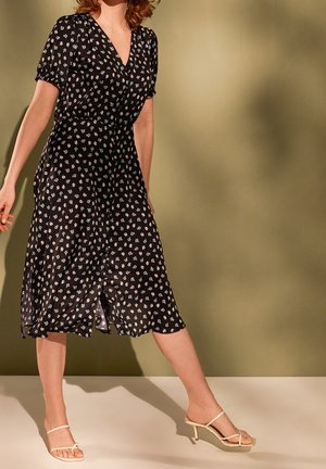 Femme portant une robe midi noire à fleurs avec manches courtes et des sandales à talons beige, debout devant un fond uni vert olive.