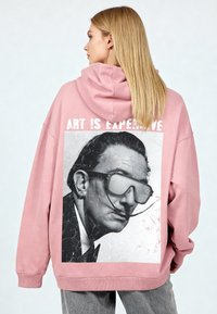 Oversized roze hoodie met een grote zwart-witte afbeelding van een man met een bril op de achterkant. De tekst luidt "ART IS EXPENSIVE." Zachte textuur.