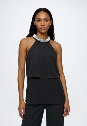 Femme aux longs cheveux foncés porte un haut noir sans manches à couches, un pantalon noir et un collier blanc épais en perles sur fond blanc.