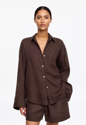 Kobieta ubrana w luźną, ciemnobrązową koszulę na guziki z kieszenią na piersi i dopasowane szorty, stojąca na tle jednolitego białego tła.