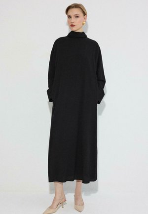 Nő hosszú, fekete, magas nyakú, hosszú ujjú ruhában, bézs hegyes orrú magassarkú cipőben és arany fülbevalóval, fehér háttér előtt állva.