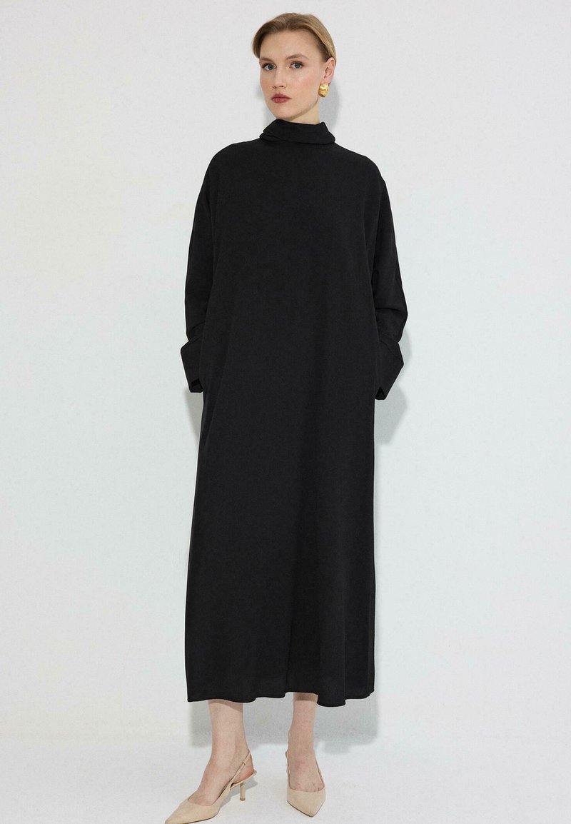 Γυναίκα που φοράει μακρύ μαύρο φόρεμα με ψηλό λαιμό και μακριά μανίκια, μπεζ παπούτσια με μυτερό δάχτυλο και χρυσά σκουλαρίκια, στέκεται μπροστά σε λευκό φόντο.