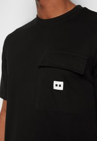 T-shirt en coton noir avec un col rond et une poche poitrine. La poche est ornée d'un logo carré blanc avec deux rectangles découpés.