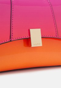 Vaaleanpunainen ja oranssi liukuvärjätty käsilaukku kiiltävällä pinnalla, jossa on kultaiset metalliosat ja kontrastiset mustat saumat. Kompakti, moderni muotoiluelementti.