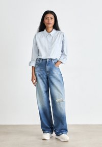 Camicia button-up azzurro chiaro con taschino sul petto, abbinata a jeans larghi di lavaggio chiaro con ginocchio strappato. Sneakers bianche completano il look.