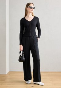 Haut noir à manches longues côtelé avec une fermeture éclair, associé à un pantalon large noir. Les accessoires comprennent un petit sac à main noir et des chaussures blanches.
