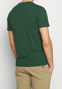 Camiseta de manga corta verde oscuro con cuello redondo. Hecha de tela suave, presenta un dobladillo recto y una textura lisa y uniforme.
