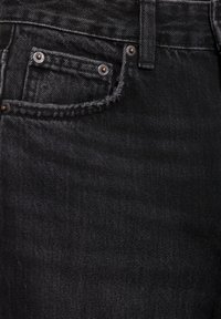 Gros plan sur un jean en denim noir montrant la poche avant, la petite poche à monnaie avec rivets et la passoire de ceinture.
