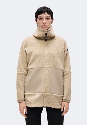 Person iført en beige fleecejakke med høj krave og lynlås foran, stående mod en ensartet lys baggrund.