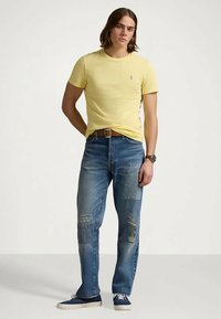 Maglietta gialla a maniche corte, jeans blue strappati con toppe, cintura marrone e scarpe da ginnastica blu. Il modello è in piedi su uno sfondo neutro.